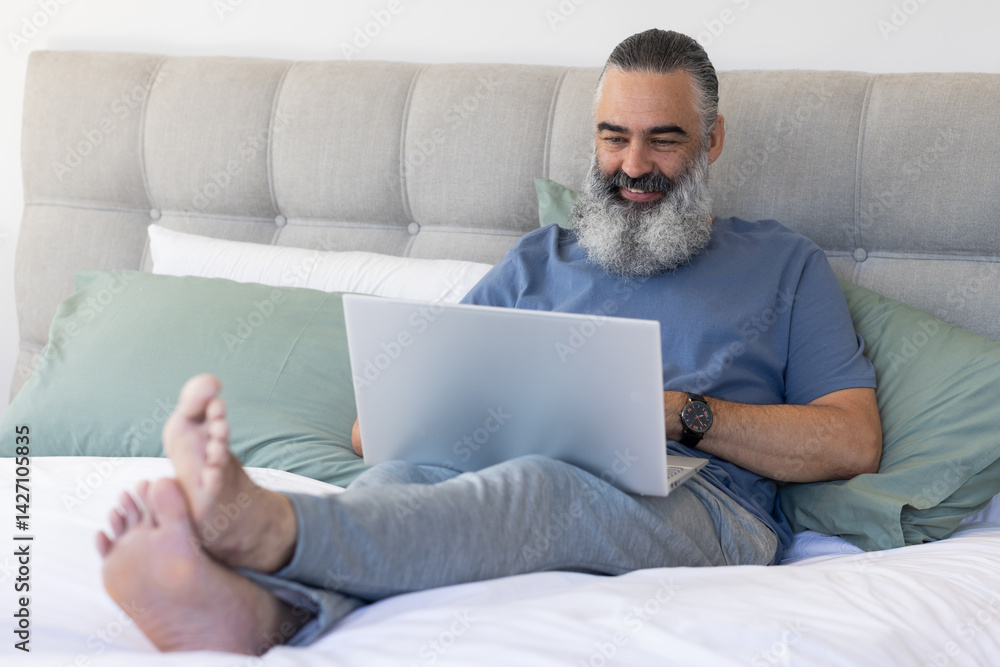 Senior man reclining on bed in bright bedroom, using silver laptop with sage green pillows