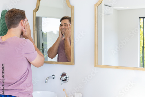 Man touching face at sink mirror in bright modern bathroom, with toothbrush and towel, copy space