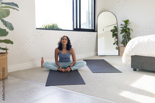 Obraz na płótnie Indian woman practicing yoga cross-legged on black mat in sunlit home gym with orange water bottle w salonie