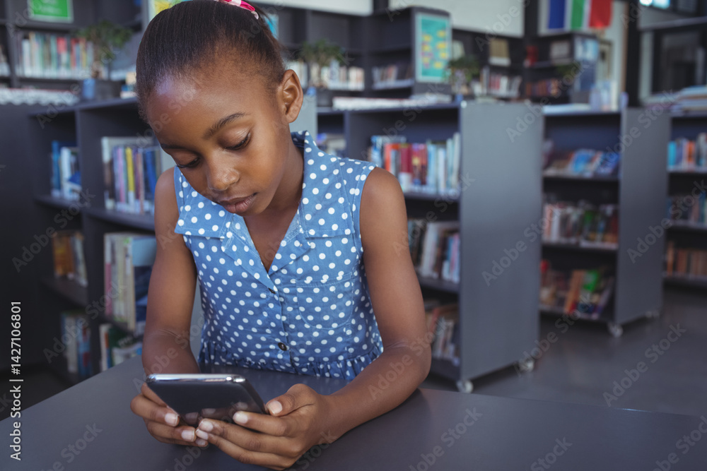African American girl child using smartphone at library table, among bookshelves and book cart
