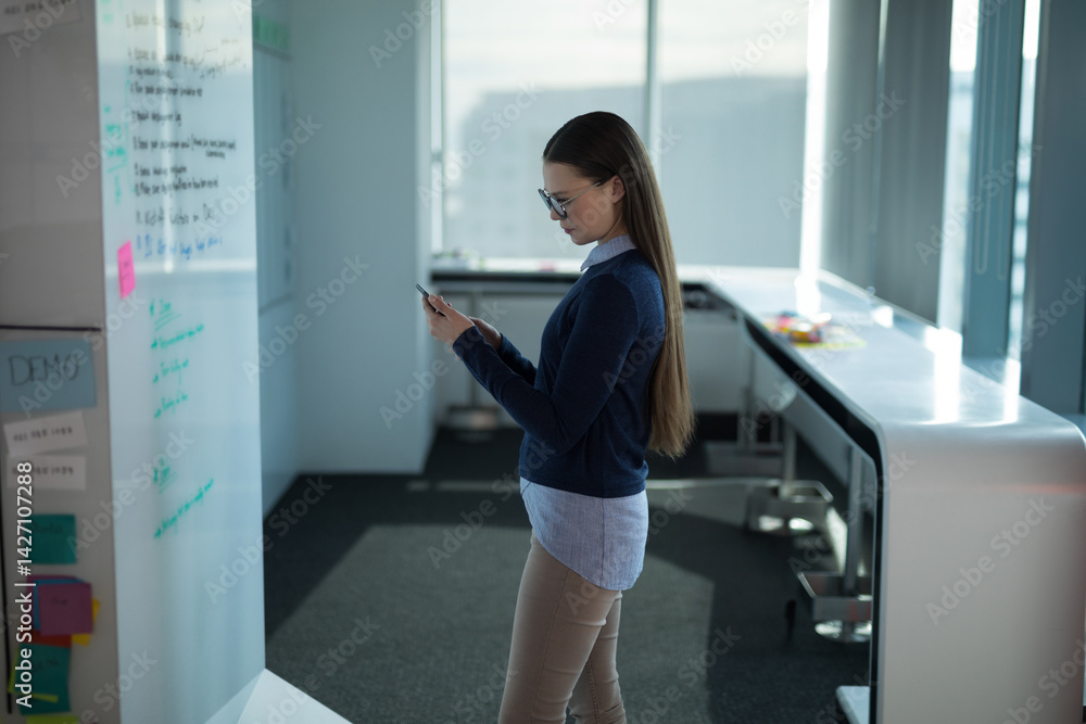 Young woman standing side-on checking smartphone in modern office near whiteboard with sticky notes