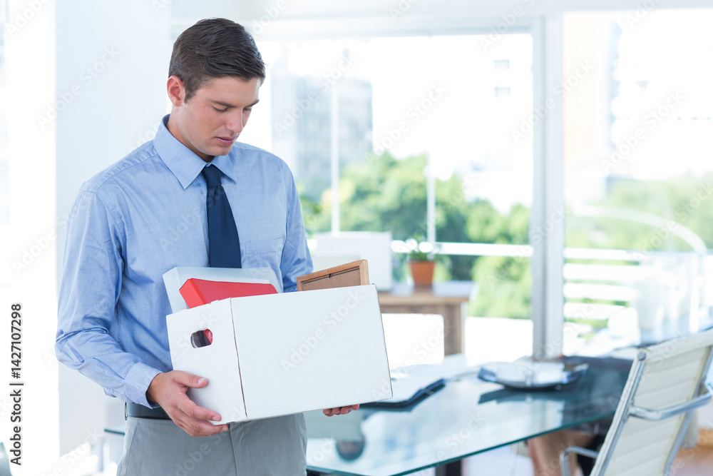 Young adult man carrying cardboard box of office items in office near windows, with glass table