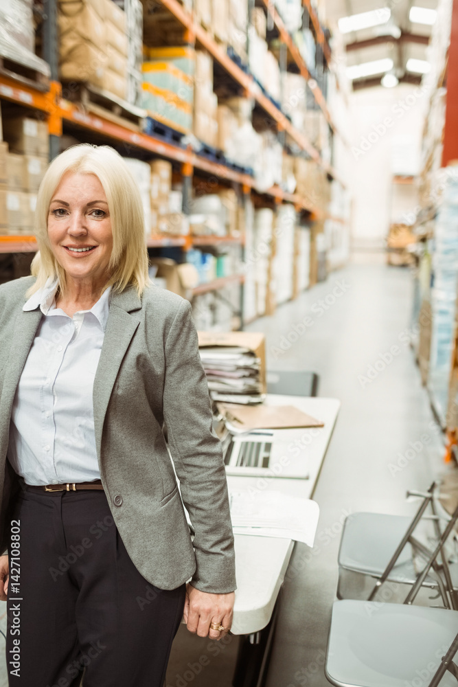 middle-aged woman standing beside work table in warehouse, checking boxes and paperwork with laptop