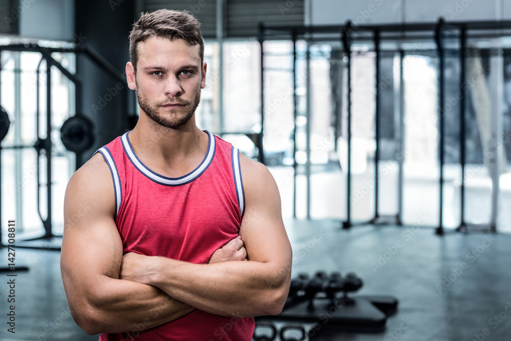 Muscular man standing with arms crossed in modern gym, with dumbbell rack and weight bench