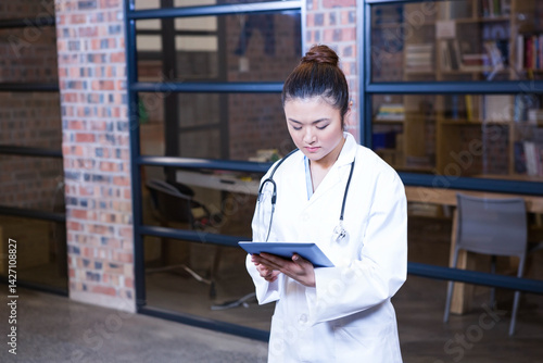 Young Asian female doctor reviewing digital tablet in medical office, with stethoscope, bookshelves