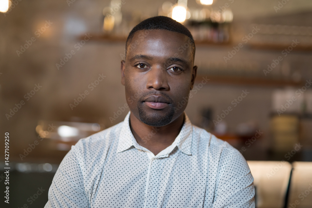 Young African American man looking into camera at cafe counter, with pastry stand, warm lights