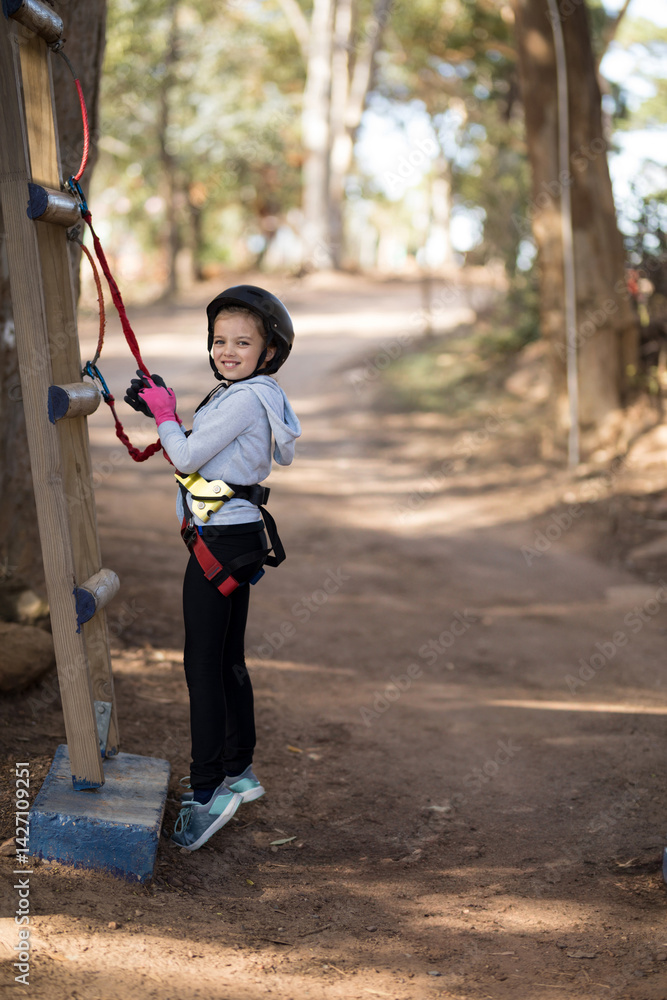 Girl climbing wooden ladder in forest adventure park, with helmet, harness and rope, copy space