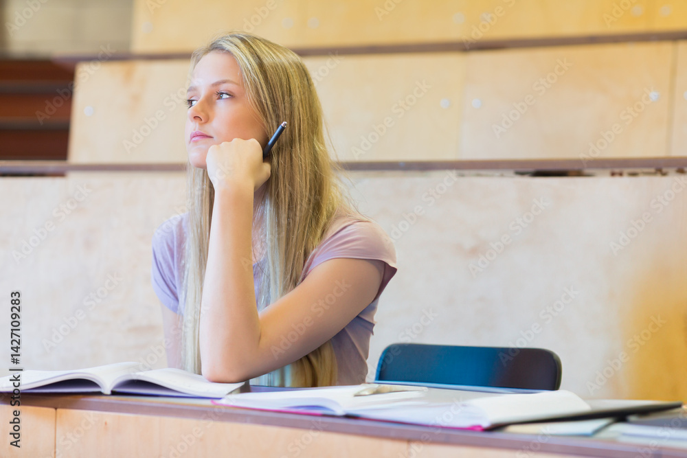 Teenage girl sitting at tiered college lecture hall desk, with pen and open textbooks and notebooks