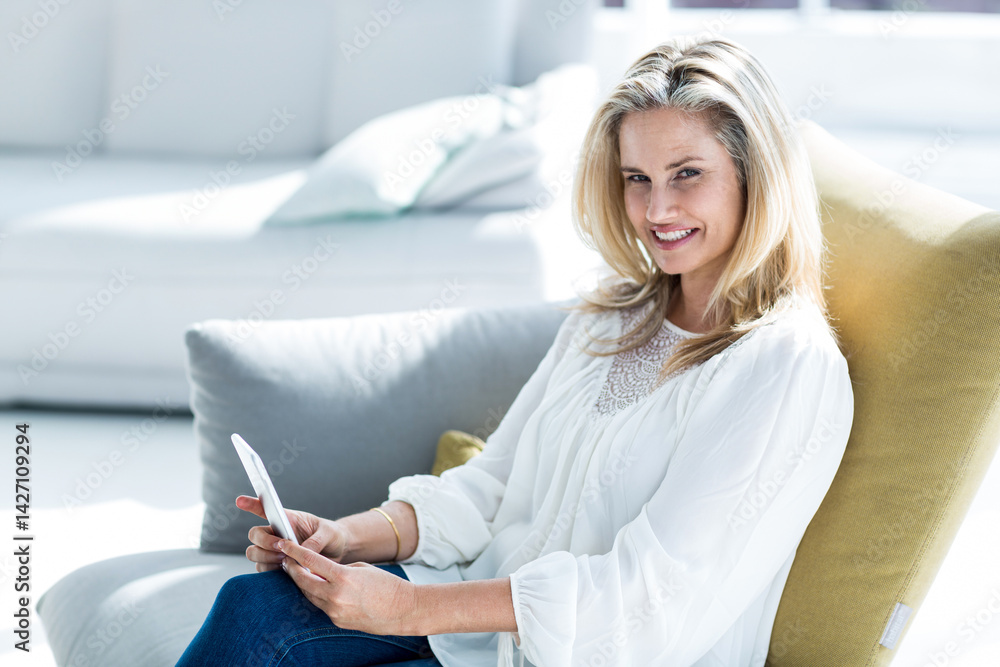 Woman sitting on grey chair with yellow cushion in living room, holding white tablet, copy space