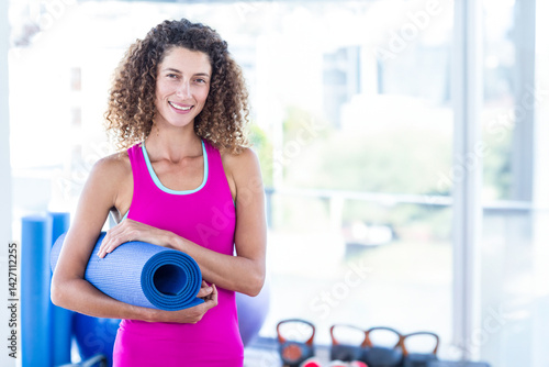 Obraz na płótnie Woman holding rolled-up yoga mat and smiling in fitness studio, with exercise ball and kettlebells w salonie