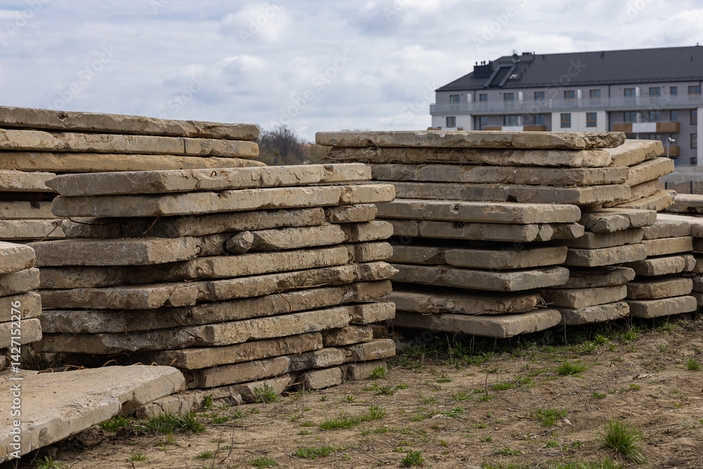 Stacks of old weathered concrete slabs at construction site with modern residential building in background on cloudy day. Urban development, recycling of materials and civil engineering Stacks of old weathered concrete slabs at construction site with modern residential building in background on cloudy day. Urban development, recycling of materials and civil engineering