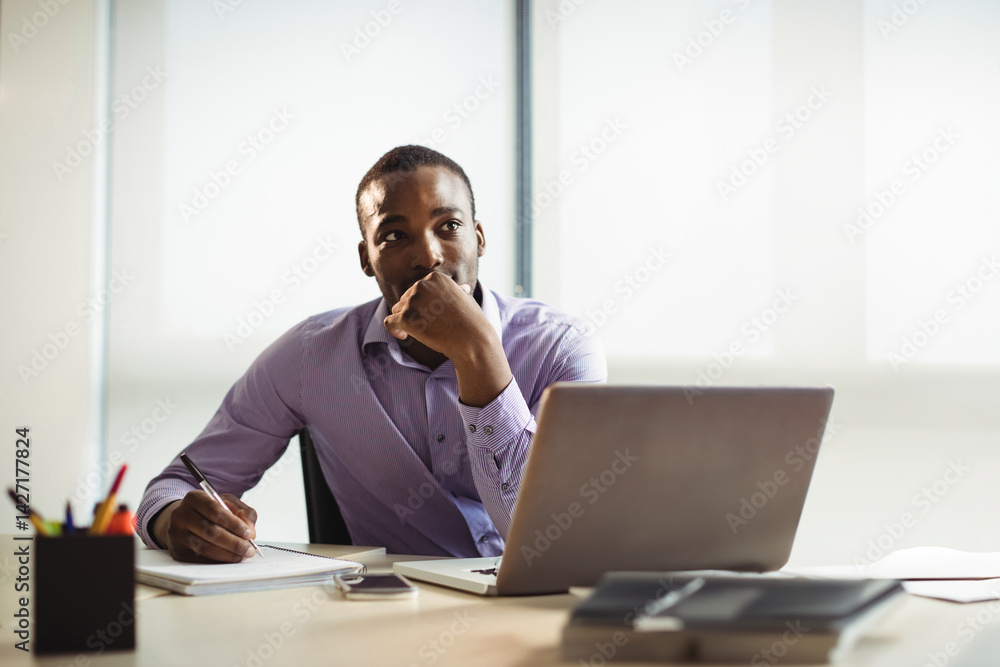 African American man writing at office desk in bright modern workspace, with laptop and notebook African American man writing at office desk in bright modern workspace, with laptop and notebook