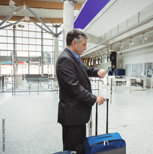 Middle-aged man checking watch and holding blue suitcase in airport hall, showing timely travel