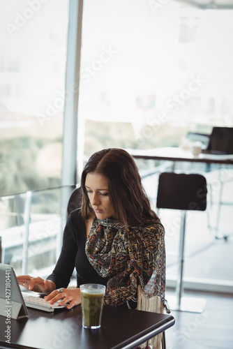 Woman typing on tablet with detachable keyboard at café table, sipping green smoothie