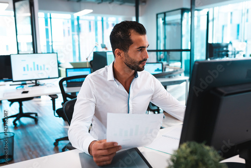 Man reviewing printed bar chart at modern open?plan office desk with computer monitor, potted plant