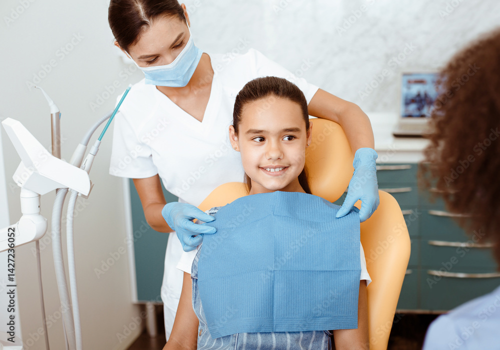 Fototapeta premium Service in modern dental clinic. Young female african american doctor looking at nurse in white coat, protective mask and gloves, dress up napkin on hispanic smiling small girl in chair, free space
