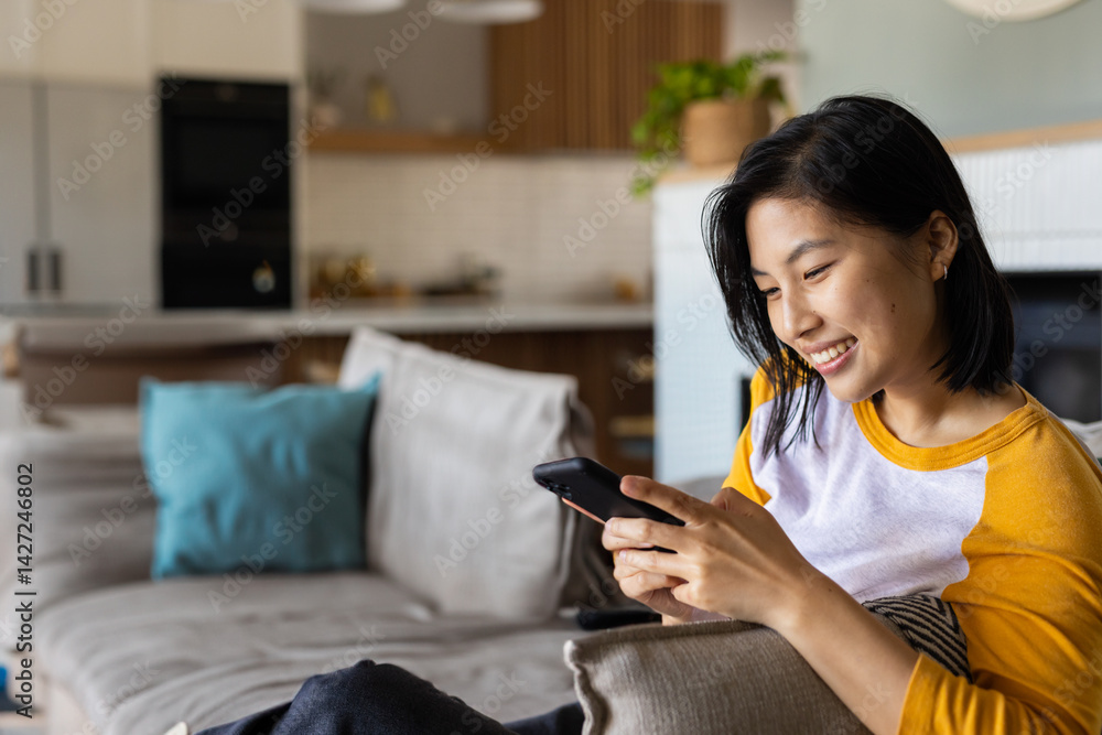 Asian woman sitting on gray sofa at home holding smartphone smiling brightly, copy space Asian woman sitting on gray sofa at home holding smartphone smiling brightly, copy space