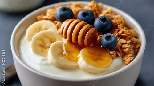 A vibrant breakfast bowl featuring banana slices, granola, and blueberries, drizzled with honey against a clean white backdrop.
