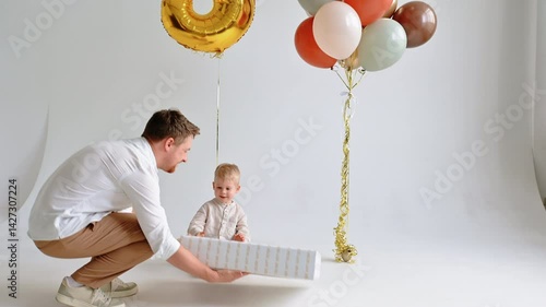 Father hands big wrapped gift to joyful toddler against bright white indoor backdrop, capturing love, surprise and family joy.
