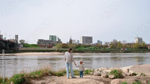 Mother and son gaze at Warsaw skyline from Vistula Riverbank on a bright summer day, blending family warmth with urban scenery.
