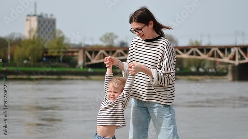Mother carries toddler along Warsaw’s Vistula riverbank, smiling together against the city skyline on a bright day, blending family warmth with urban scenery.
