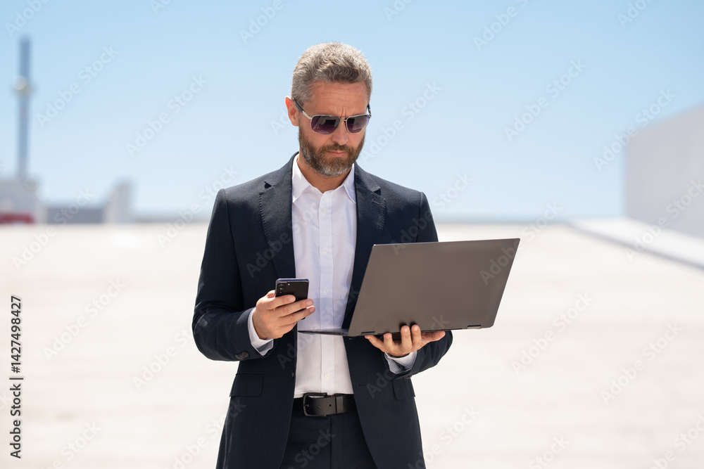 Businessman with phone and laptop outdoors. Mature businessman online business. Businessman with laptop. Business online. businessman checking data on laptop outside. Business man in suit work online.