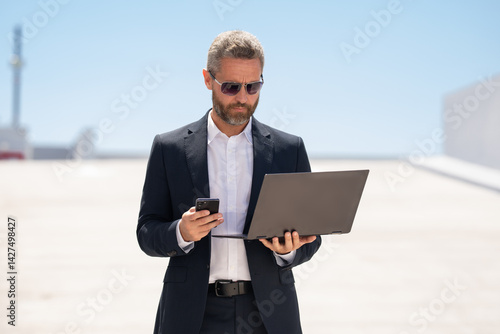 Obraz na płótnie Businessman with phone and laptop outdoors. Mature businessman online business. Businessman with laptop. Business online. businessman checking data on laptop outside. Business man in suit work online. w salonie