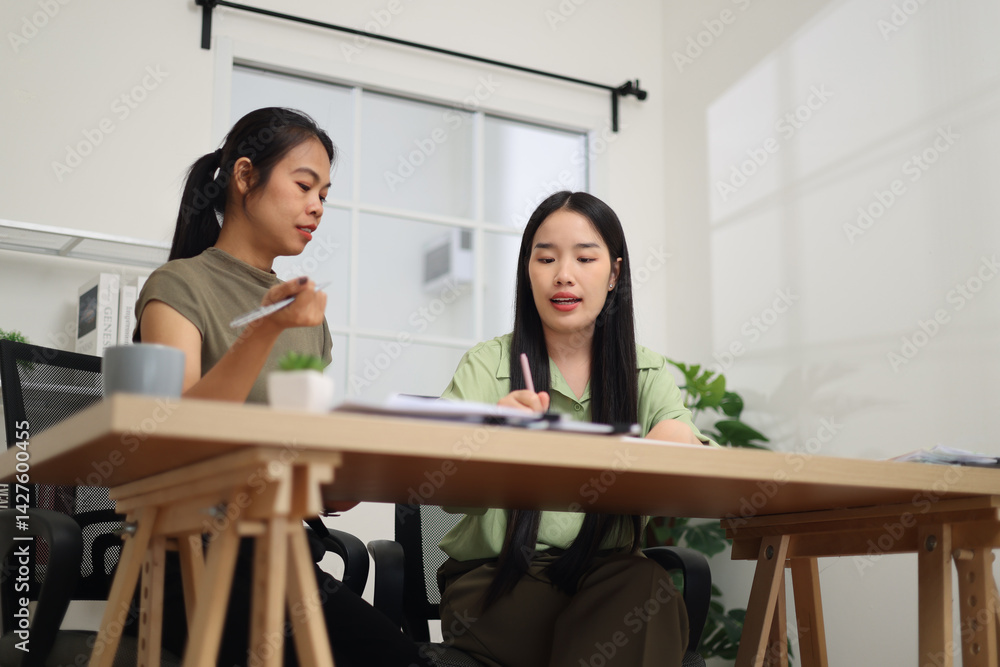 Low angle view shot, Two female colleagues are sitting together at desk, reviewing documents and discussing work in the office.