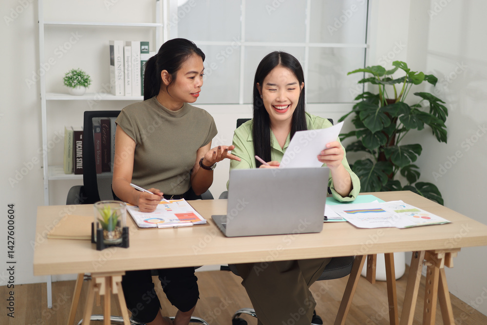 Portrait of Two female colleagues sit together at a desk, holding documents, discussing a business plan and exchanging ideas in a modern workplace.