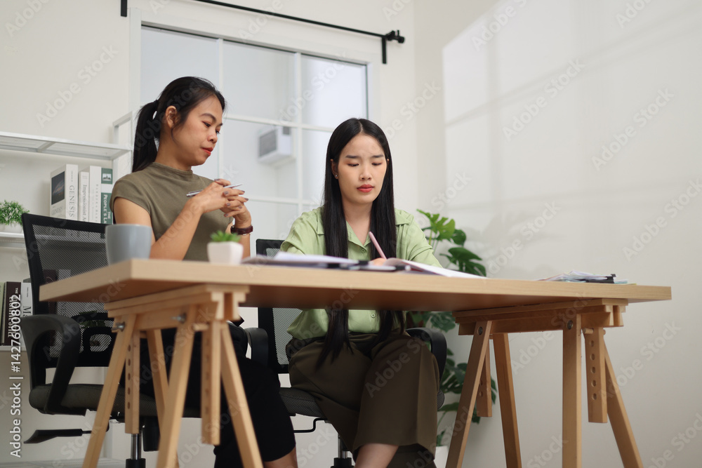 Low angle view shot, Two female colleagues are sitting together at desk, reviewing documents and discussing work in the office.