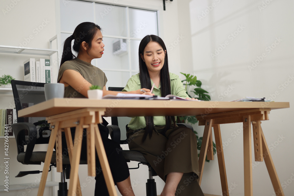 Low angle view shot, Two female colleagues are sitting together at desk, reviewing documents and discussing work in the office.
