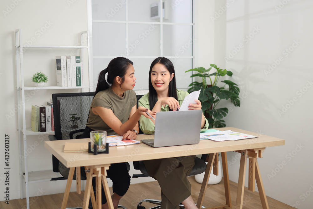 Portrait of Two female colleagues sit together at a desk, holding documents, discussing a business plan and exchanging ideas in a modern workplace.