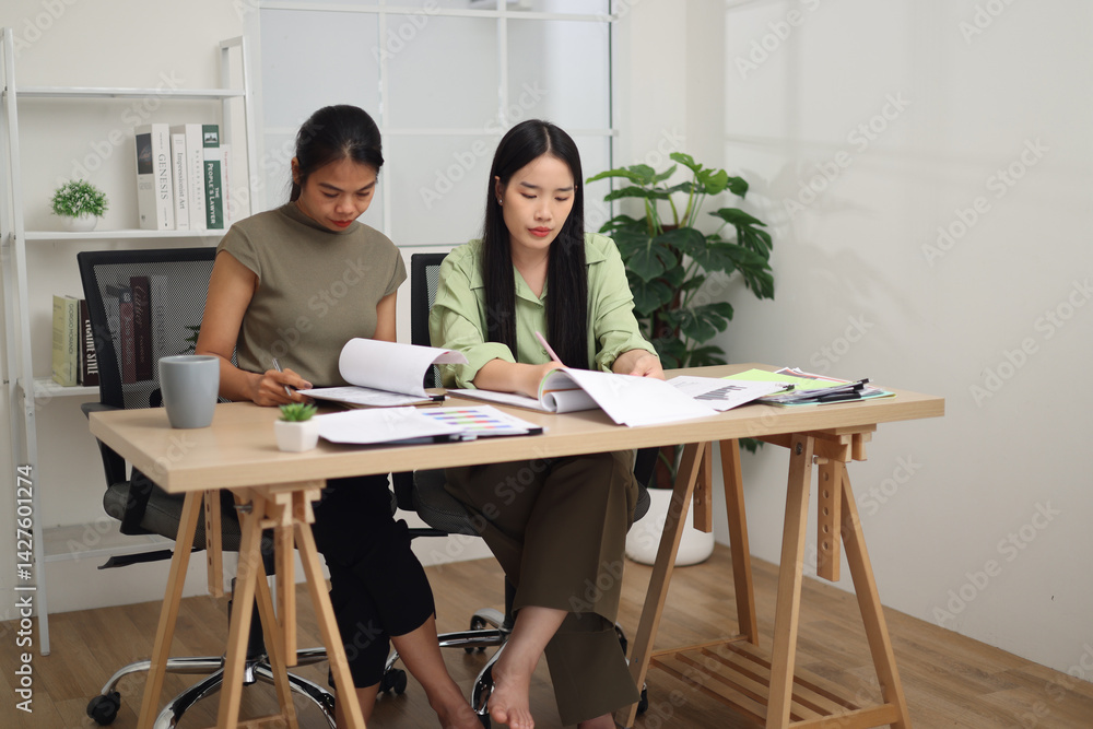 Portrait of Two female colleagues are sitting together at desk, reviewing documents and discussing work in the office. Portrait of Two female colleagues are sitting together at desk, reviewing documents and discussing work in the office.