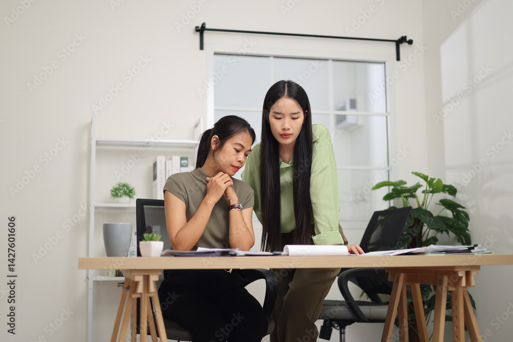 Portrait of Two female colleagues are working together in an office. One stands explaining the work, while the other sits and listens with interest.