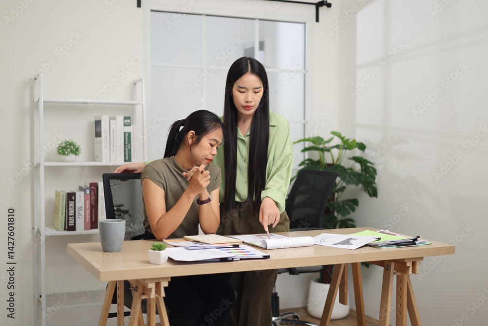 Portrait of Two female colleagues are working together in an office. One stands explaining the work, while the other sits and listens with interest.