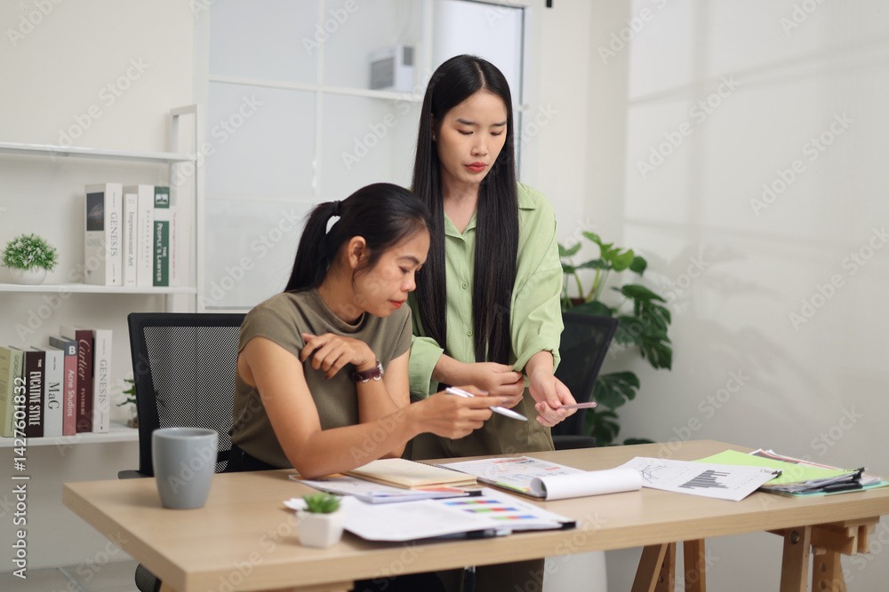 Portrait of Two female colleagues are working together in an office. A young woman is standing, talking and exchanging ideas with another colleague who is sitting.