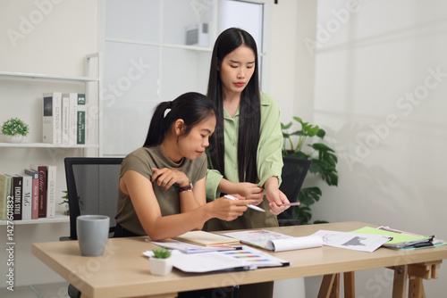 Obraz na płótnie Portrait of Two female colleagues are working together in an office. A young woman is standing, talking and exchanging ideas with another colleague who is sitting. w salonie