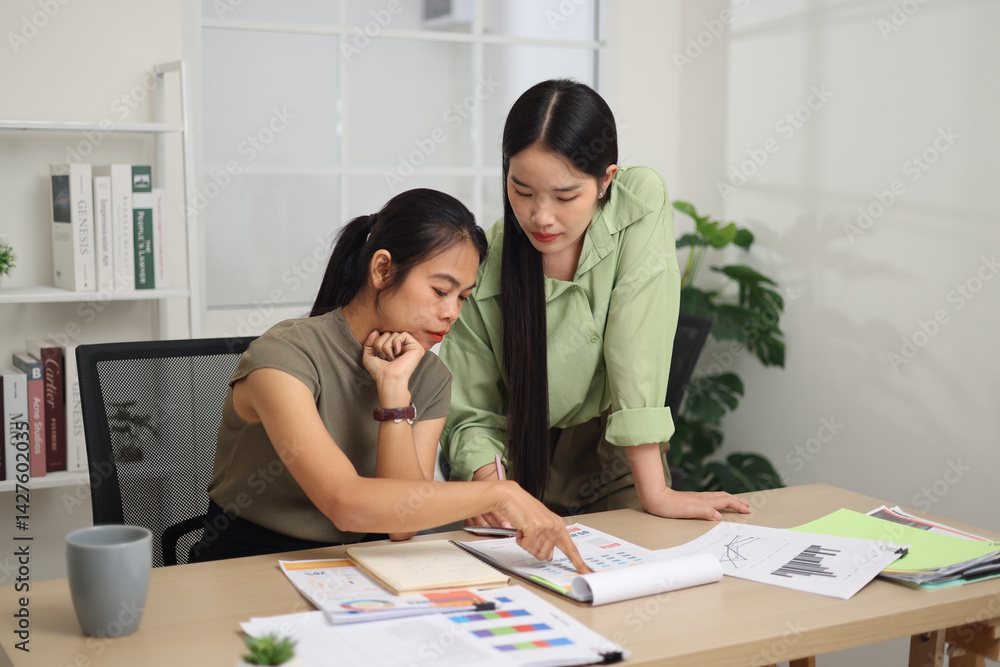 Portrait of Two female colleagues are working together in an office. One stands explaining the work, while the other sits and listens with interest.