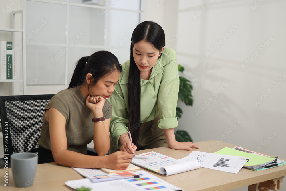 Portrait of Two female colleagues are working together in an office. A young woman is standing, talking and exchanging ideas with another colleague who is sitting.