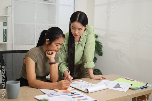 Portrait of Two female colleagues are working together in an office. A young woman is standing, talking and exchanging ideas with another colleague who is sitting.