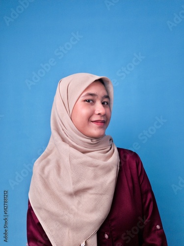 Portrait of Smiling Southeast Asian Muslim Woman in Hijab Looking to the Side Facing Forward Against Blue Background