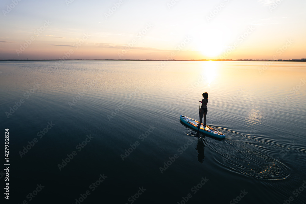 a young woman stands on a board with a paddle and swims on a beautiful lake. Girl on sup board with beautiful colored sunrise background top view from the throne