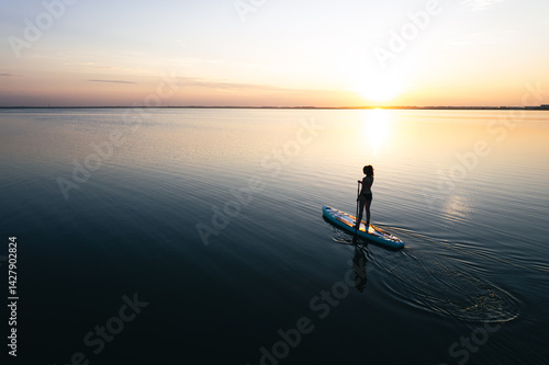 Obraz na płótnie a young woman stands on a board with a paddle and swims on a beautiful lake. Girl on sup board with beautiful colored sunrise background top view from the throne w salonie