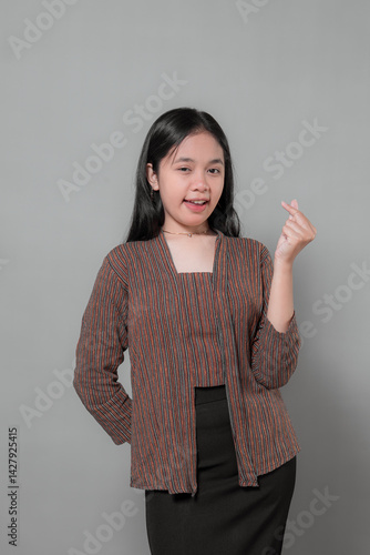 Asian woman in traditional striped lurik blouse gives a soft smile while making a Korean finger heart gesture, conveying warmth, friendliness, and subtle charm in studio setting.