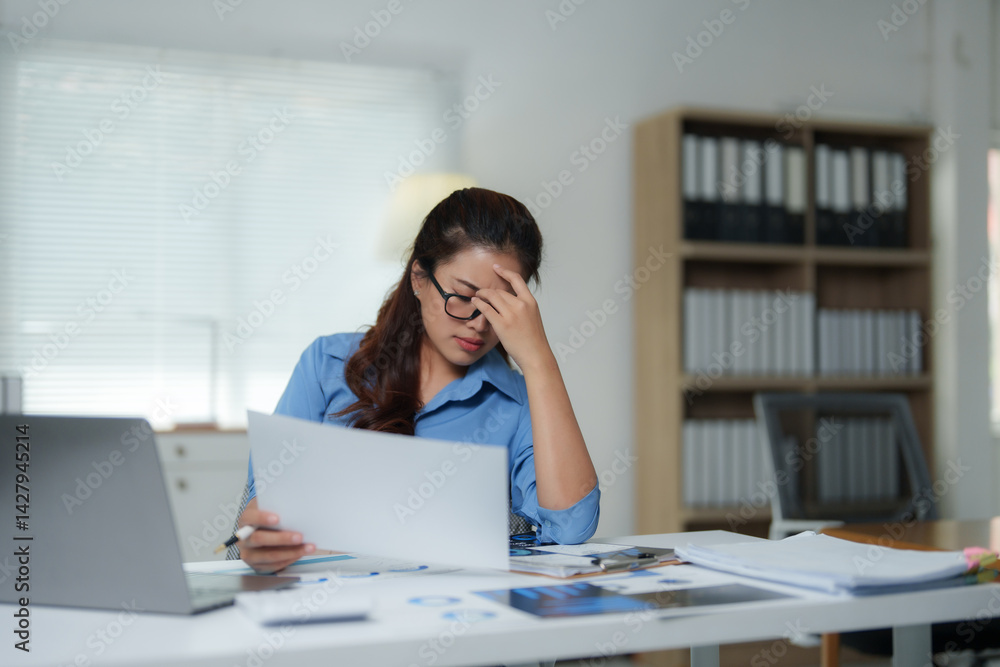 Young asian businesswoman sitting at her desk in modern office with laptop and documents, she is having a bad headache and feeling tired and stressed Young asian businesswoman sitting at her desk in modern office with laptop and documents, she is having a bad headache and feeling tired and stressed