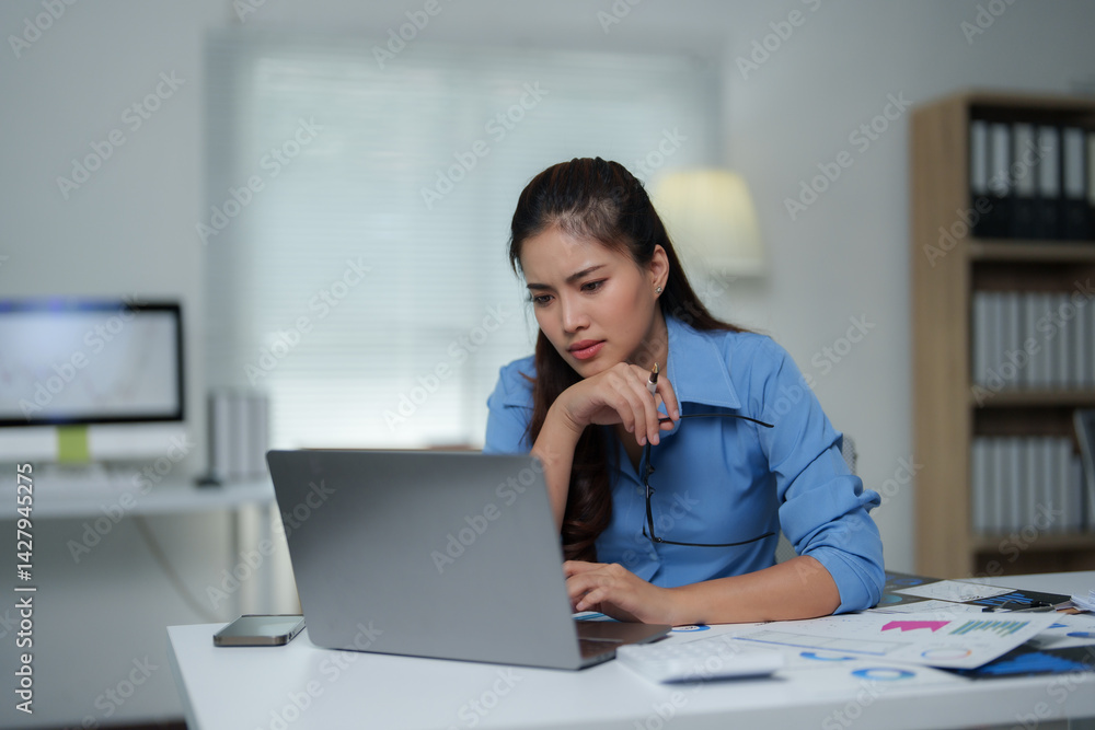Businesswoman working on laptop with focused expression, holding eyeglasses and analyzing financial data in modern office, with desktop computer and documents on table Businesswoman working on laptop with focused expression, holding eyeglasses and analyzing financial data in modern office, with desktop computer and documents on table