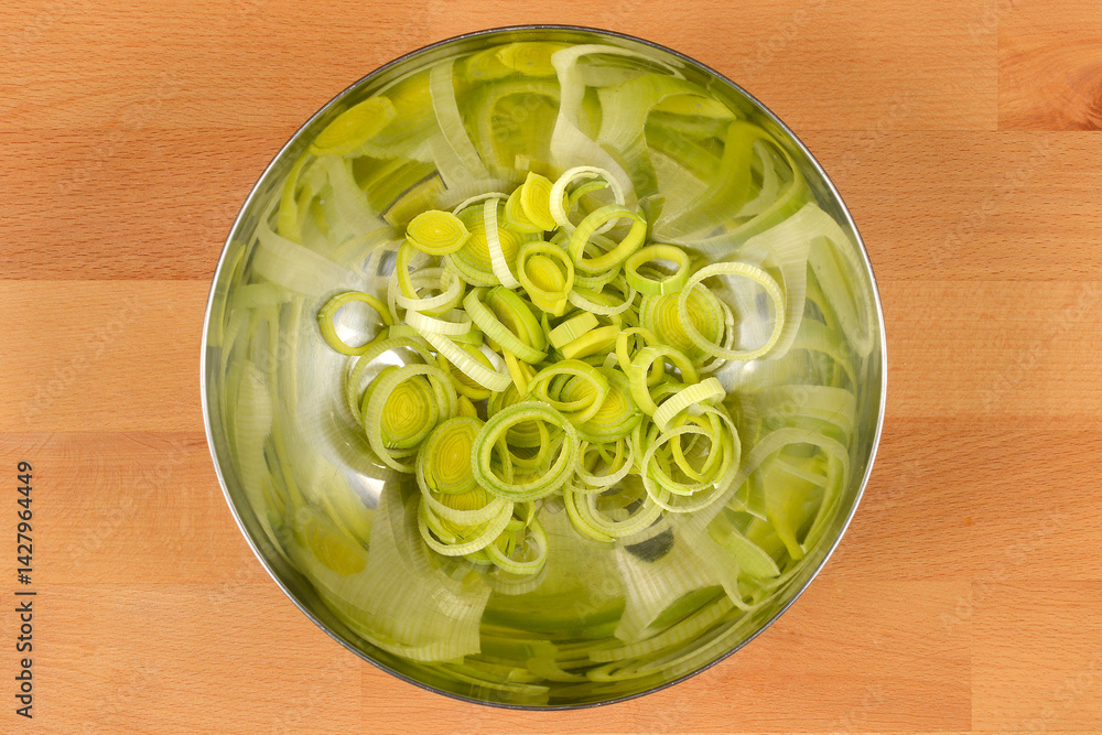A silver bowl contains a pile of sliced light green and white leeks on a light brown wooden surface