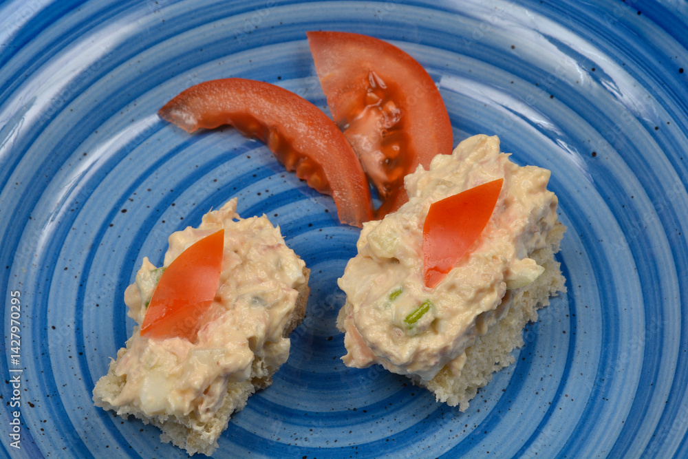 Two small bread slices topped with tuna, cream cheese, egg, caper, onion, and mustard spread are garnished with tomato on a blue plate