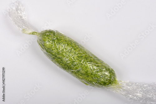 A log of vibrant green wild garlic (Allium ursinum) butter is tightly wrapped in clear plastic wrap on a light surface, captured from a slightly elevated angle
