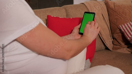 Close-up of a pregnant woman sitting on a sofa and holding a green screen phone in her hands. 